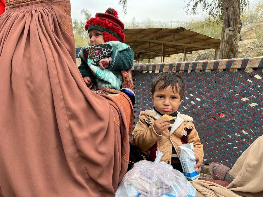 A woman in a burq holds baby in a woolly hat as an older child looks into the camera eating a WFP biscuit